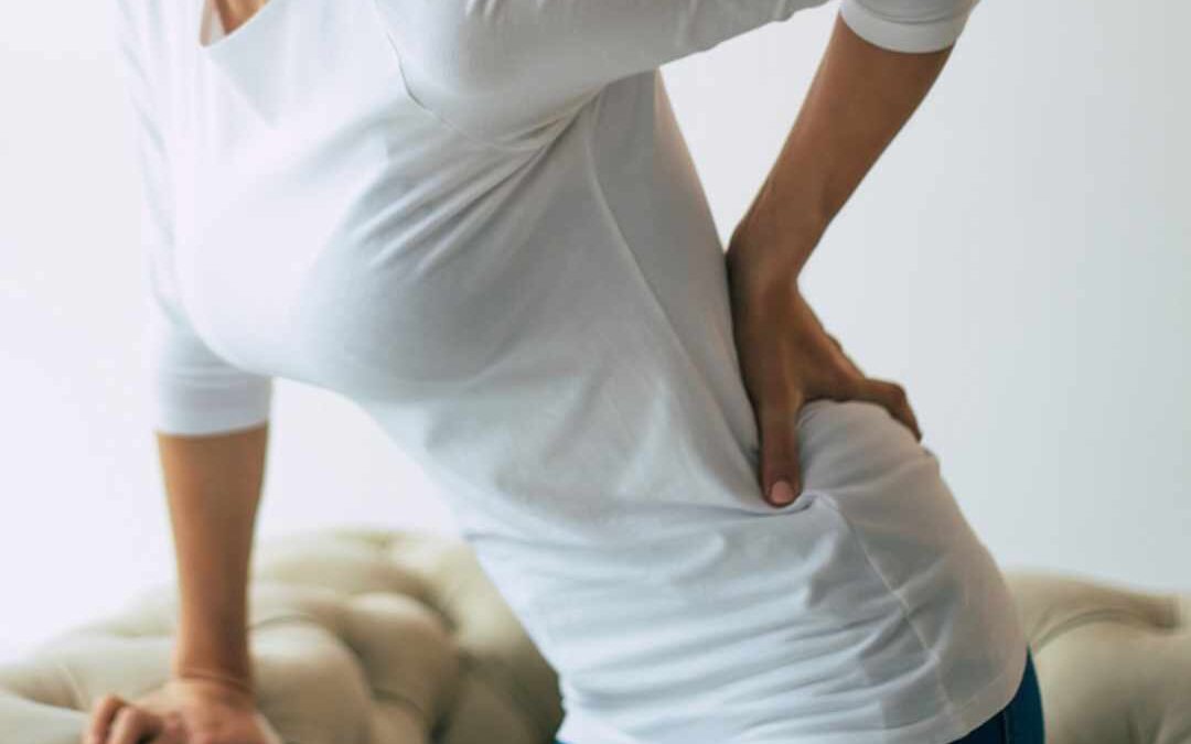 Kvinde iført hvid trøje og jeans står ved en sofa, holder sig på den nederste del af ryggen med én hånd og støtter sig til sofaen med den anden, som tegn på smerter i lænd og hofte.