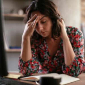Kvinde med hovedpine sidder ved skrivebord med hånden på panden – illustrerer stress og migræne.