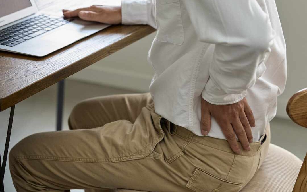 Person sidder på stol foran computer og holder sig på lænden – illustrerer smerter i muskler og led.