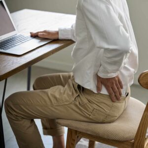 Person sidder på stol foran computer og holder sig på lænden – illustrerer smerter i muskler og led.