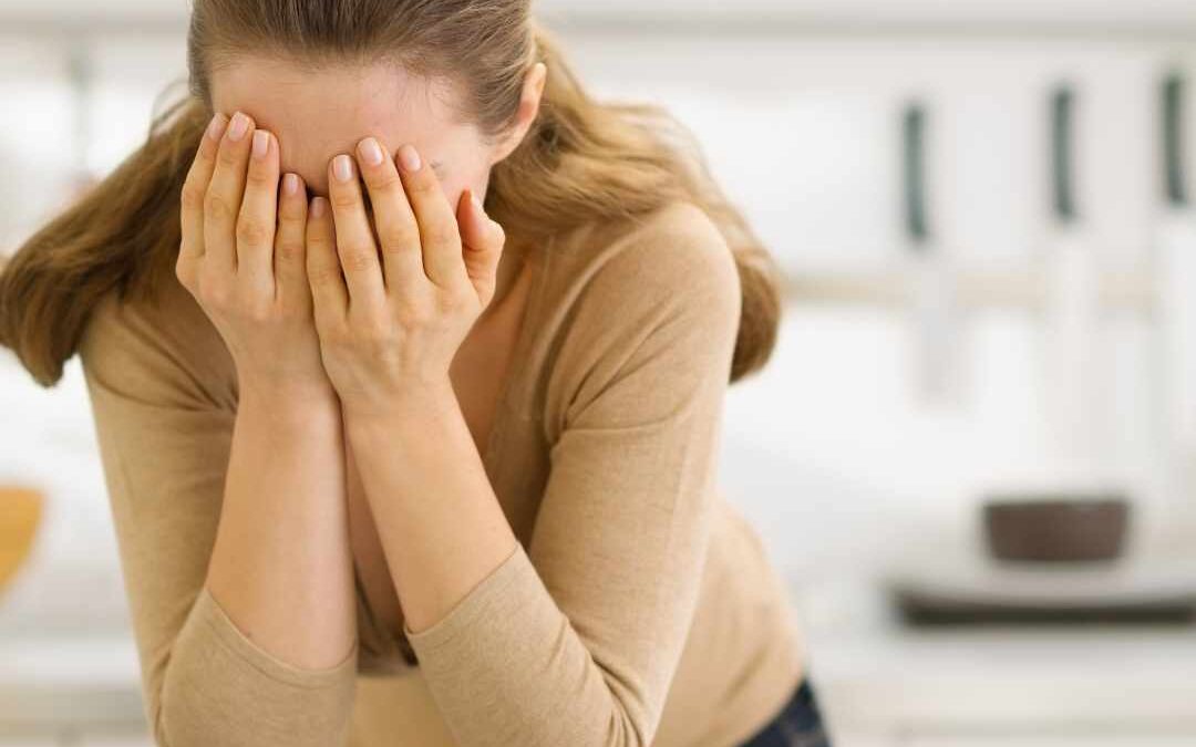 Kvinde står foroverbøjet med hænderne for ansigtet i et lyst køkkenmiljø, hvilket signalerer tegn på stress eller følelsesmæssig overbelastning.