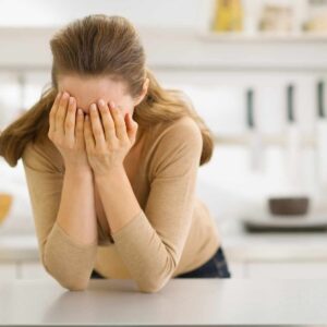 Kvinde står foroverbøjet med hænderne for ansigtet i et lyst køkkenmiljø, hvilket signalerer tegn på stress eller følelsesmæssig overbelastning.