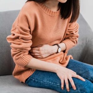 Ung kvinde sidder foroverbøjet og presser hænderne mod maven med tydelige spændinger i maven, ofte relateret til ubehag, stress eller fordøjelsesbesvær.
